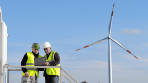 Two workers in hard hats reviewing a laptop beside a wind turbine